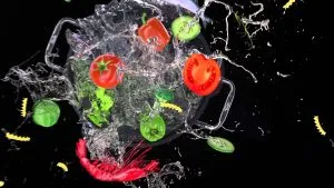 various food items such as tomatoes, peppers, cucumbers, shrimp, and pasta being splashed with water in a pot